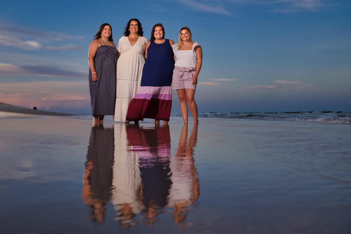 Four women standing on the beach during sunset, smiling, with their reflections visible in the wet sand.