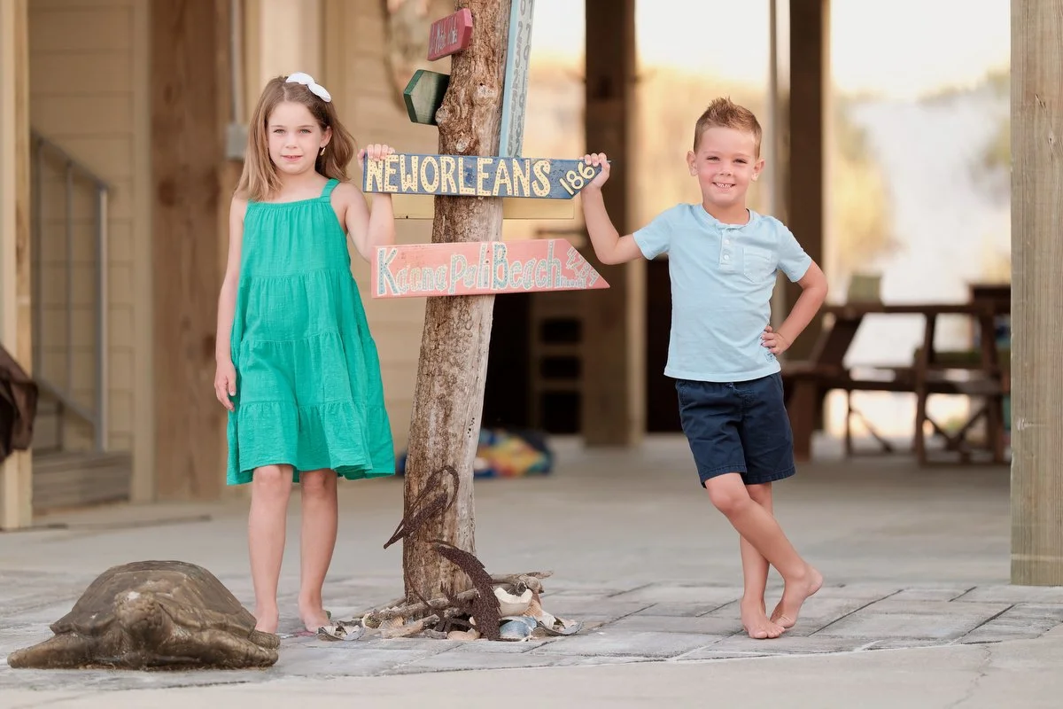 A young girl in a green dress and a young boy in a light blue shirt and navy shorts stand on a boardwalk near a signpost with signs for New Orleans and Kiawah Palmetto Beach. They are smiling and holding the signpost. There is a turtle figure on the 