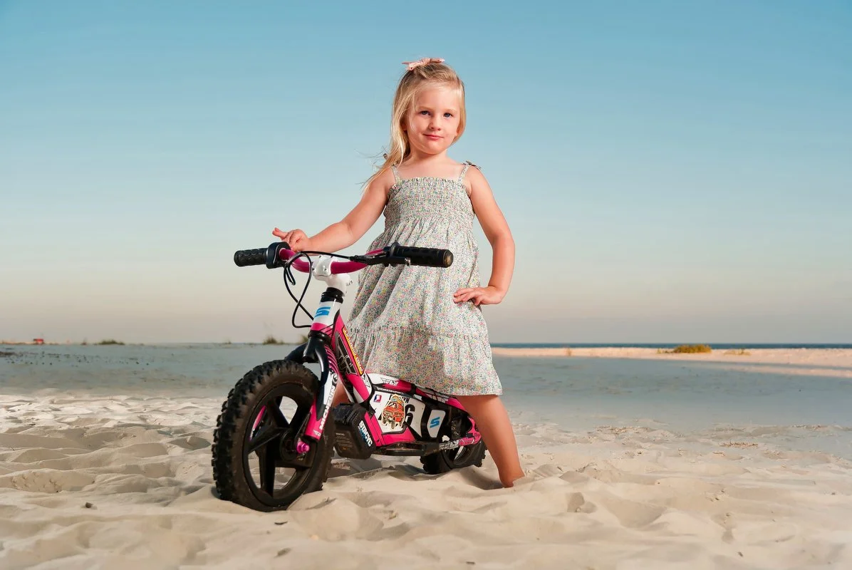 A young girl in a summer dress standing on a sandy beach with one foot on a pink and black children's bicycle. She has blonde hair with a bow and is looking at the camera with a slight smile under a clear blue sky.