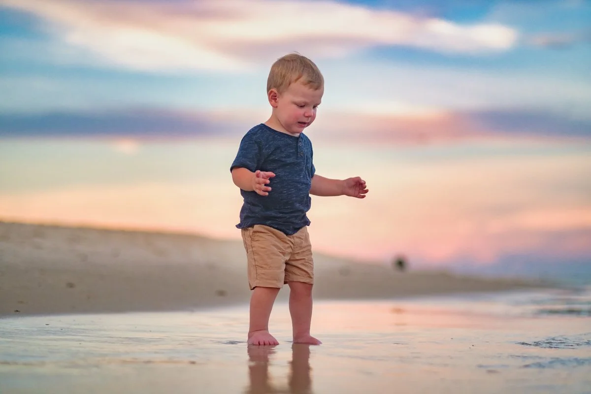 Young boy in a blue shirt and tan shorts standing at the shoreline, playing in shallow water during sunset.