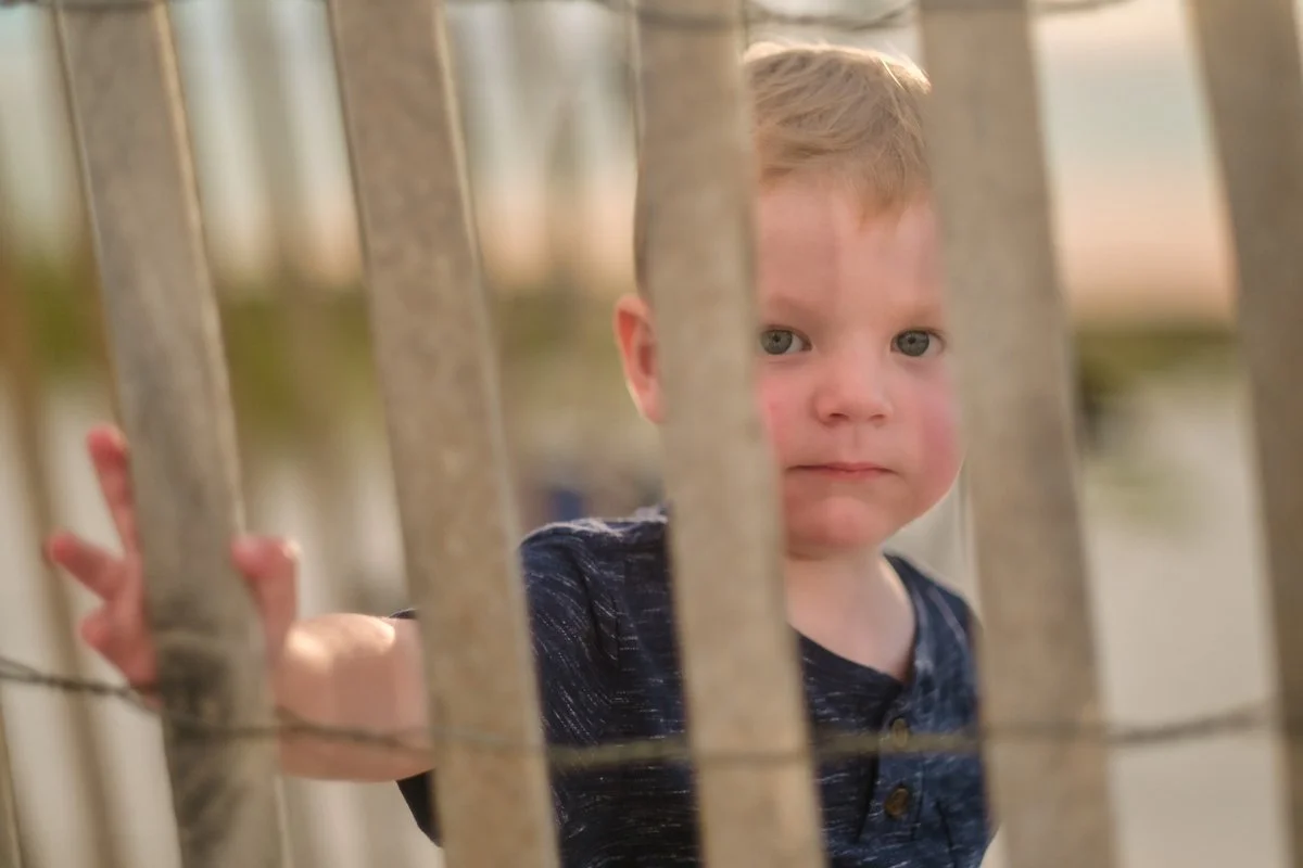 A young boy with blond hair and blue eyes looking through metal bars of a fence.