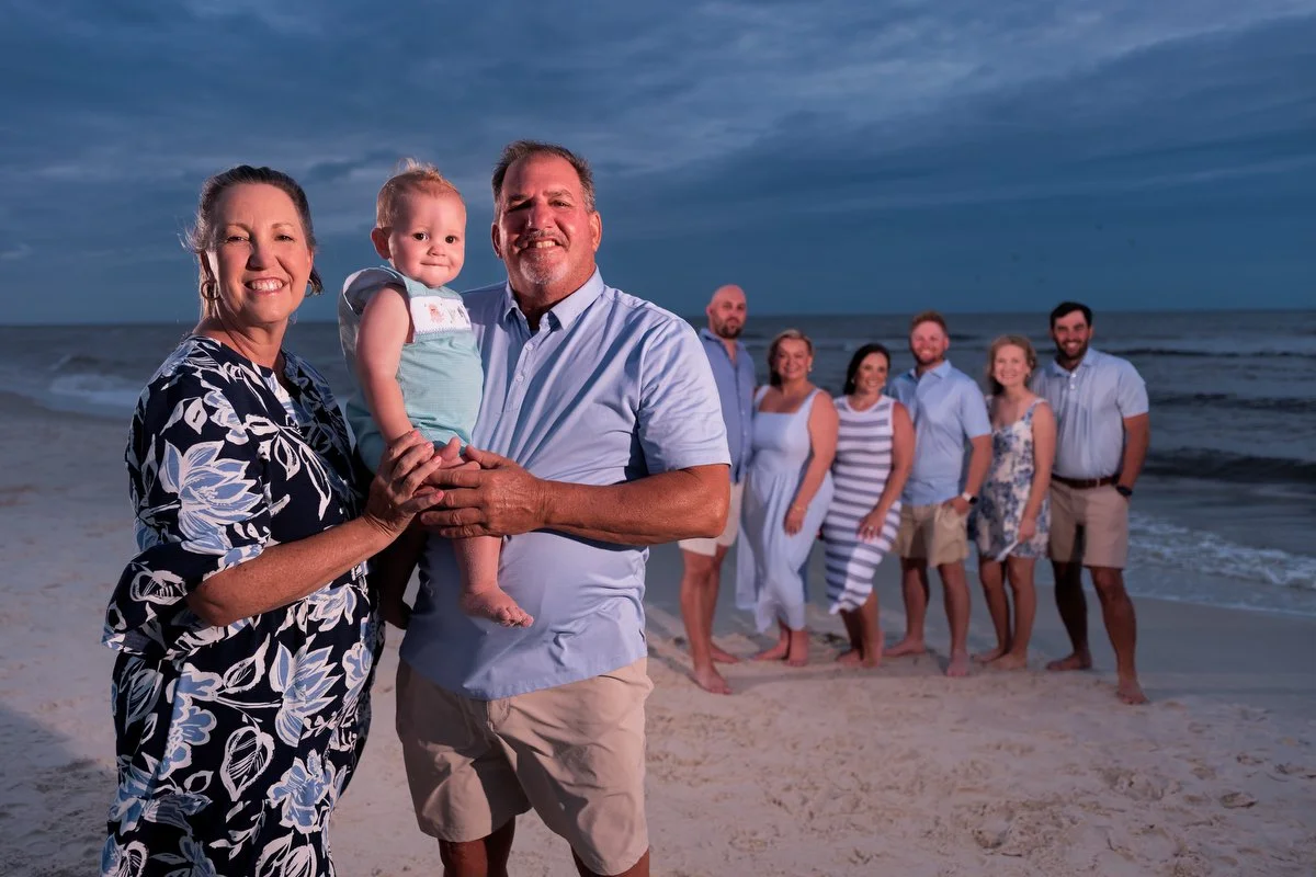 Family standing on a beach during sunset, with the ocean and dark cloudy sky in the background. The group includes a woman, a man holding a young girl, and five other adults standing in a line behind them, all dressed in casual summer clothing.