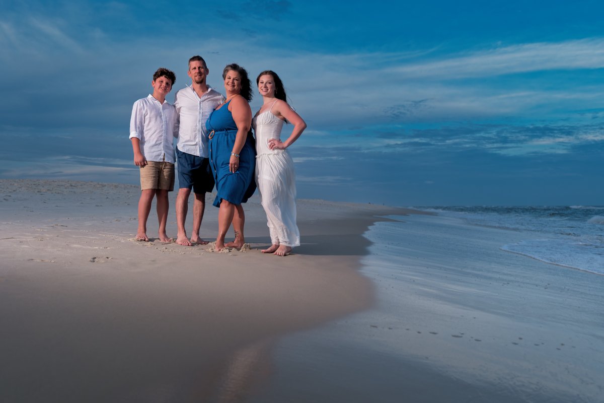 Group of four people standing on a sandy beach near the shoreline during daytime, with blue sky and clouds in the background.