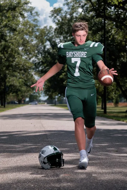 Young football player in a green Bayshore jersey with the number 7, holding a football, running on a tree-lined street with a helmet on the ground nearby.