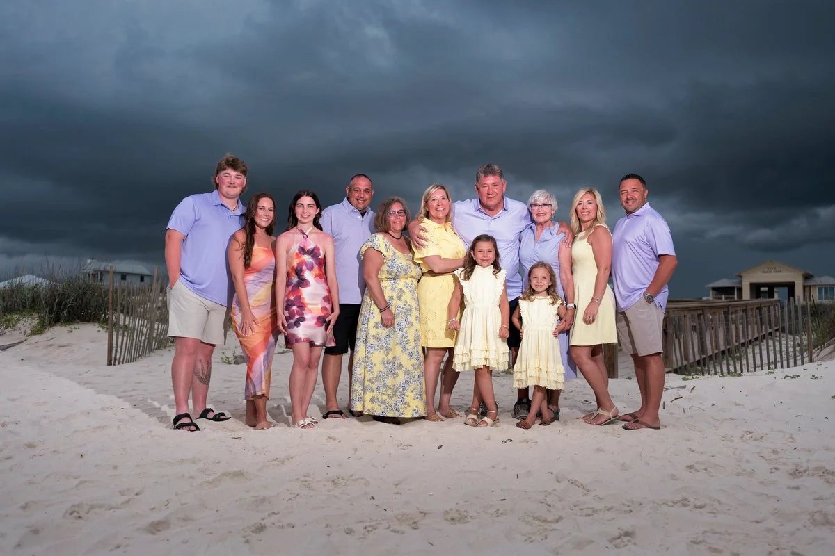 A large family group standing on a sandy beach with dark storm clouds overhead. The group includes adults and children dressed in summer clothing, some smiling at the camera at Kiva Dunes in Gulf Shores.