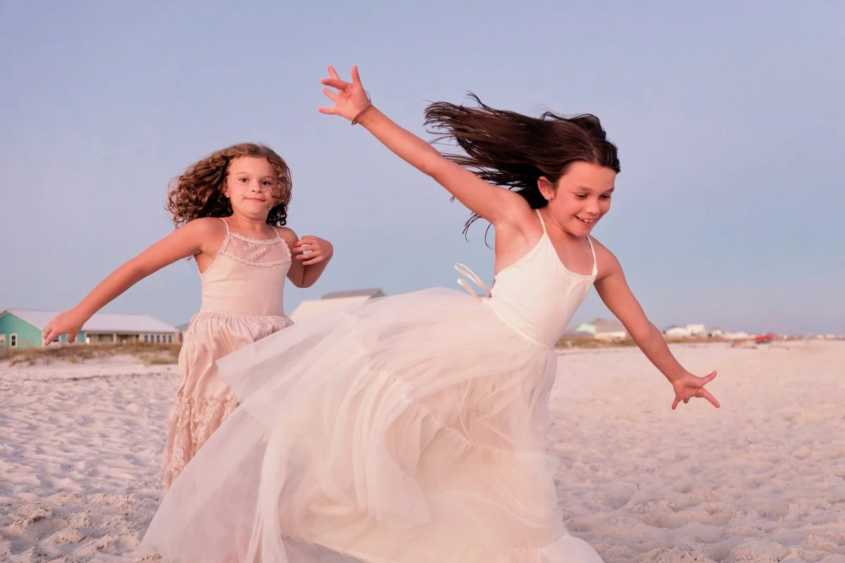 Two young girls in dresses playing on the beach in Gulf Shores, one with long dark hair and the other with curly light brown hair, during daytime.
