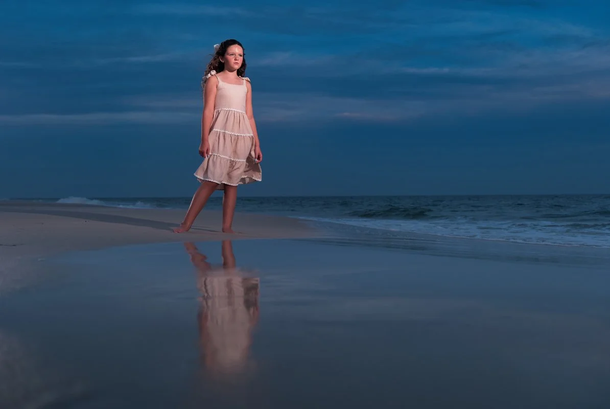 A young girl in a light pink dress standing barefoot on the wet sand at the beach, with her reflection visible in a shallow pool of water, during dusk or dawn with a cloudy sky.