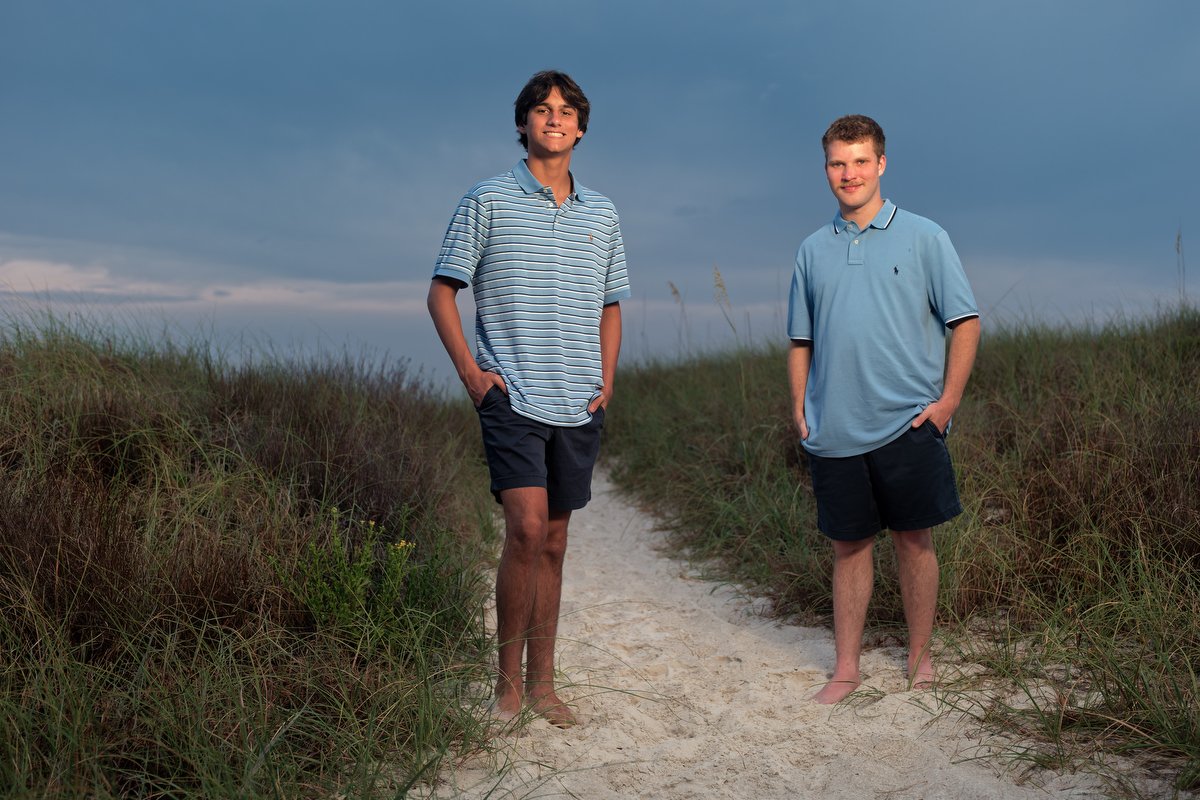 Two young men standing barefoot on a sandy beach path with grass on either side at the Island House Hotel in Orange Beach, under a cloudy sky using off camera flash. 