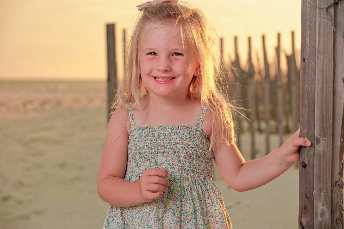 A smiling young girl with blonde hair, wearing a colorful floral dress, standing near a wooden post at the beach during sunset.