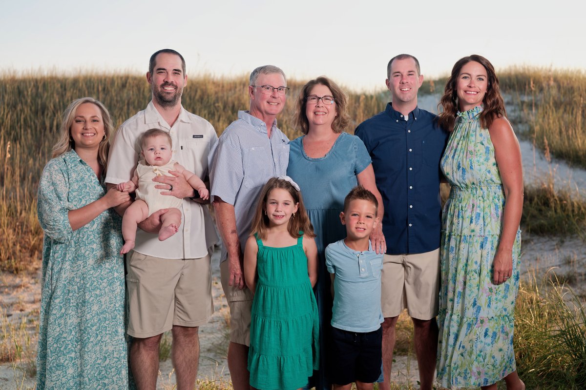 Family group photo on a beach with sand dunes and grass in the background, consisting of adults and children smiling for the camera.