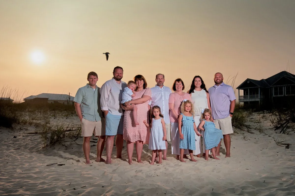 Family group photo on the beach at sunset with seven adults and four children, some in pastel dresses, and a bird flying in the sky.