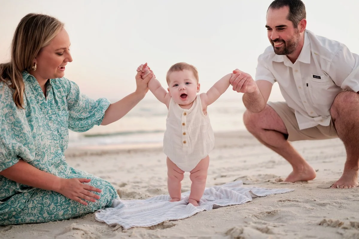 A happy family at the beach, with a woman and man holding a toddler's hands as the toddler learns to stand on the sand.