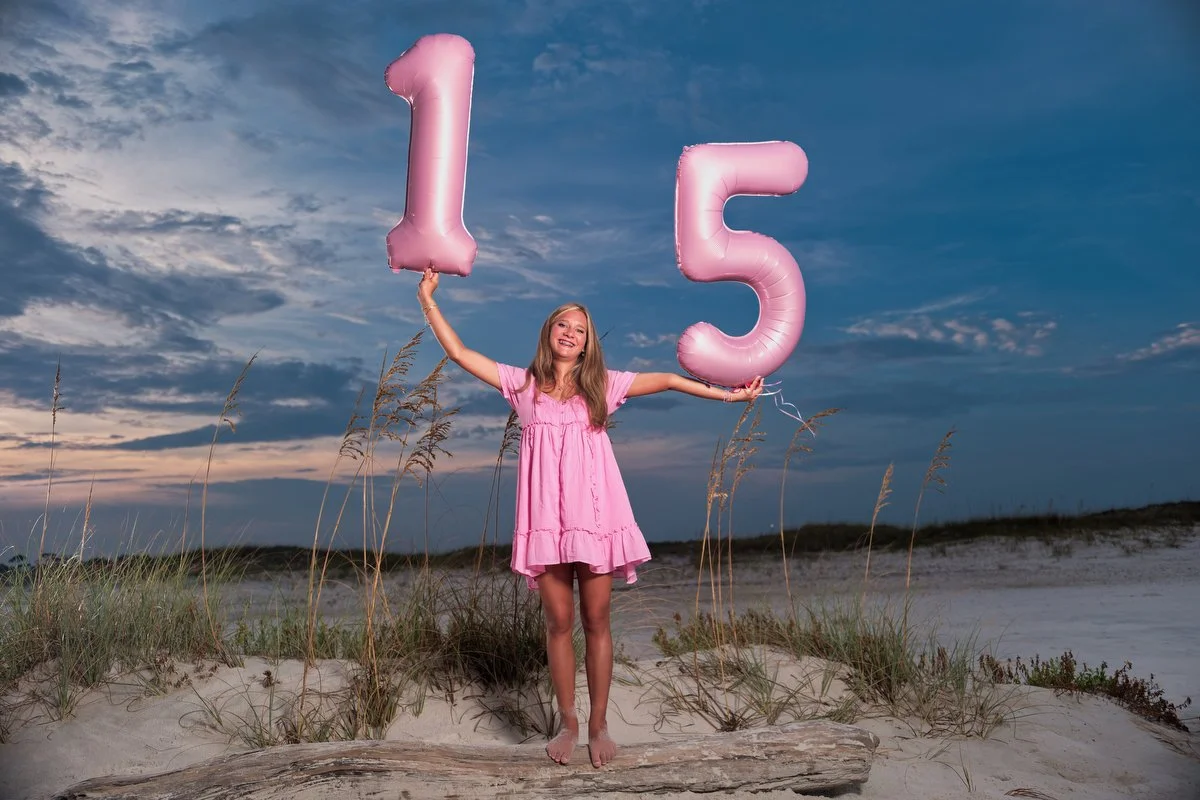 A young girl in a pink dress on a beach holding pink balloons shaped as the numbers one and five, celebrating her 15th birthday in Perdido Key using off camera flash. 