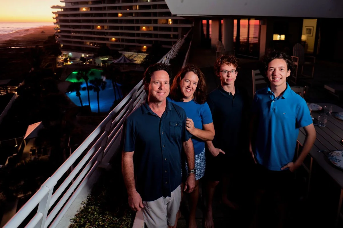 A family of four smiling on a balcony overlooking a resort pool and the ocean at sunset.