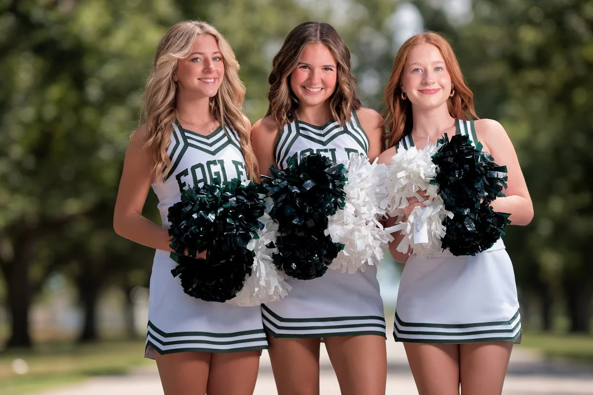 Three young women cheerleaders standing outdoors on a sunny day, holding black and white pom-poms, wearing matching green and white cheerleading uniforms with 'EAGLE' written on the front.