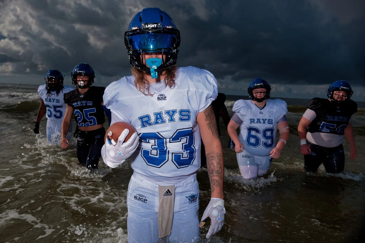 A group of football players from the Rays team wading into the ocean, wearing helmets and jerseys, with dark cloudy skies overhead, taken at the Gulf Shores Public Beach using off camera flash