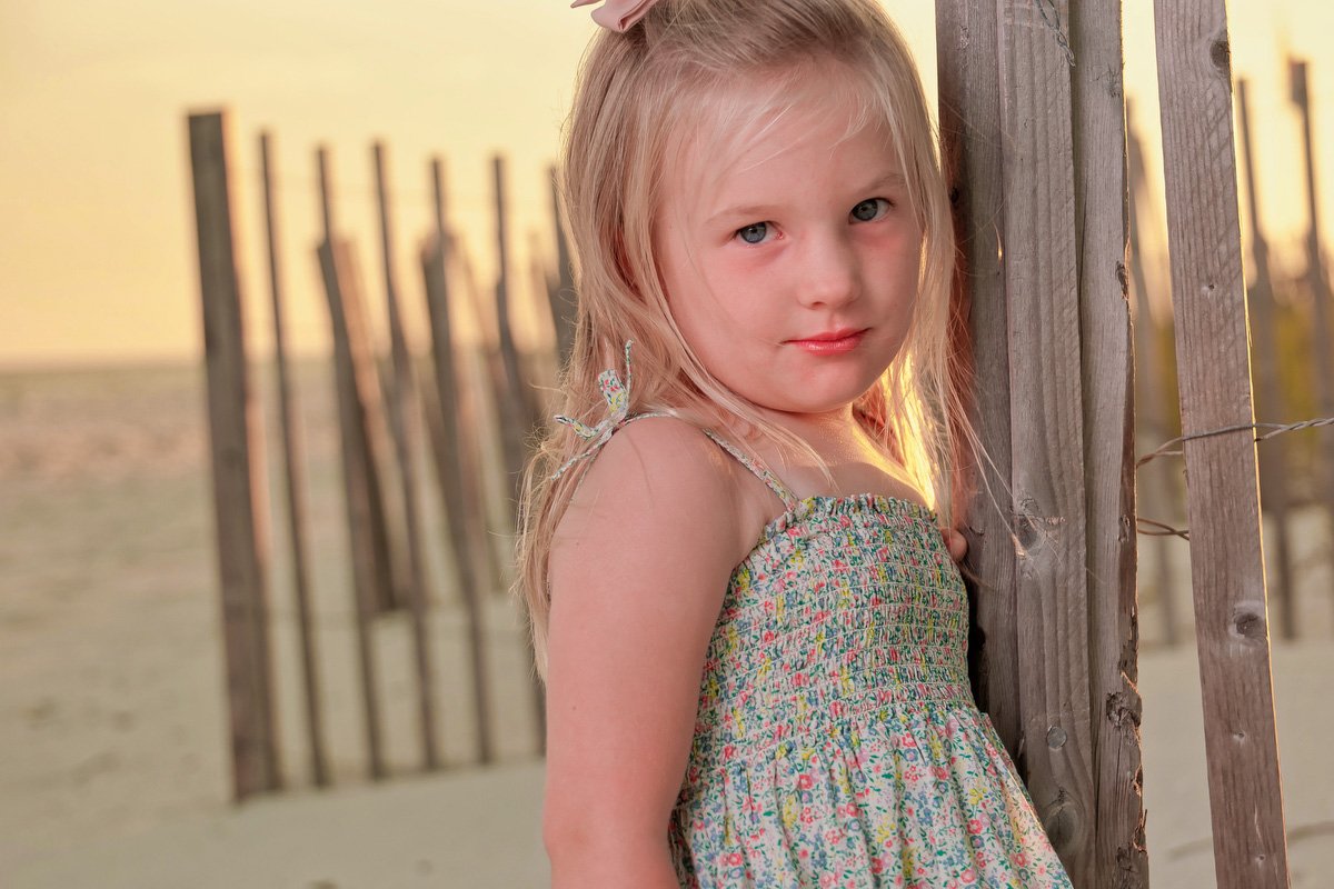 A young girl with blonde hair, blue eyes, and a pink bow in her hair, leaning against a wooden fence on a beach at sunset, wearing a colorful floral dress.