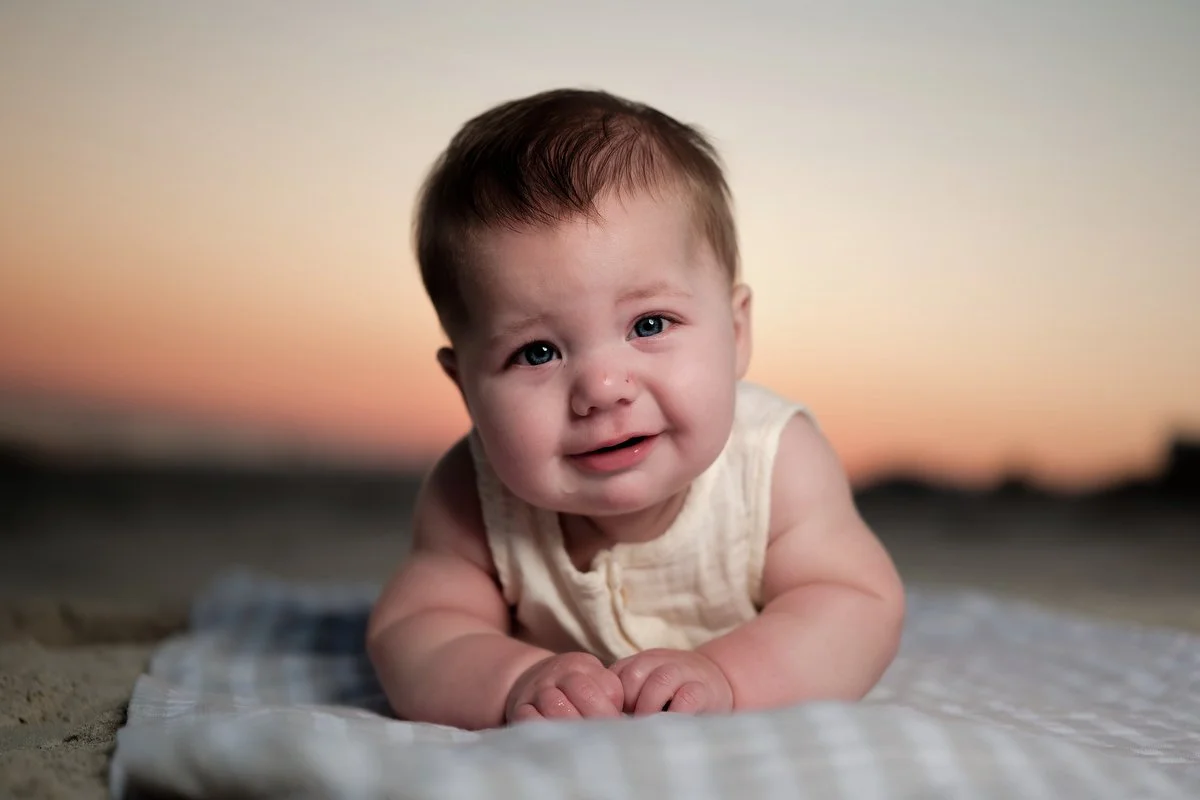 A baby with blue eyes and brown hair is crawling on a blanket on the sand at sunset.