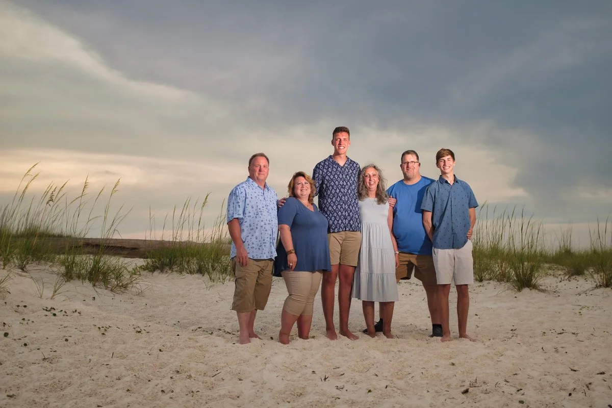 Family of six standing on a sandy beach with tall grasses, cloudy sky in the background, smiling and posing for a photo.