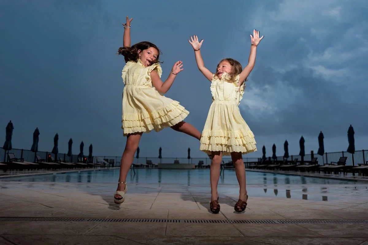 Two young girls in yellow dresses jumping and playing near a pool outdoors during the evening or dusk, with a cloudy sky in the background in Fort Morgan.