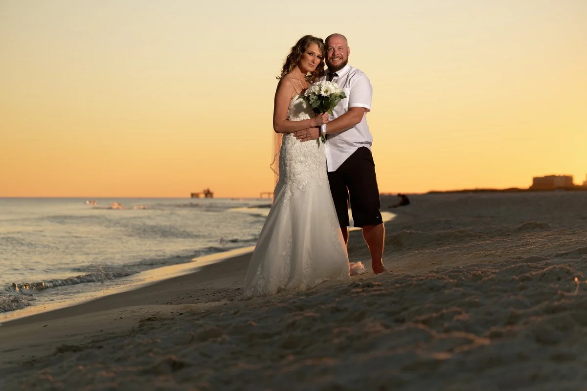 A bride and groom standing on a beach during sunset, with the bride holding a bouquet of flowers in Gulf Shores
