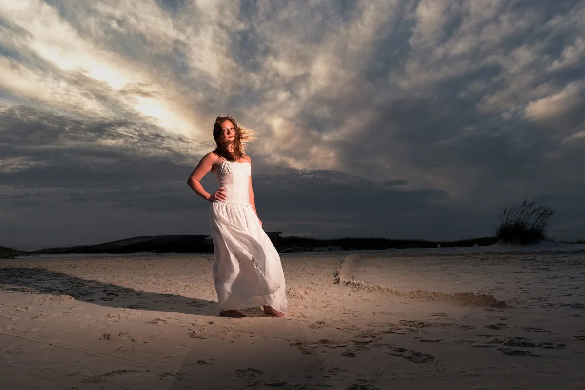A woman in a white dress standing on a sandy beach during sunset with dramatic clouds in the sky with off camera flash used to give the photo more color and depth