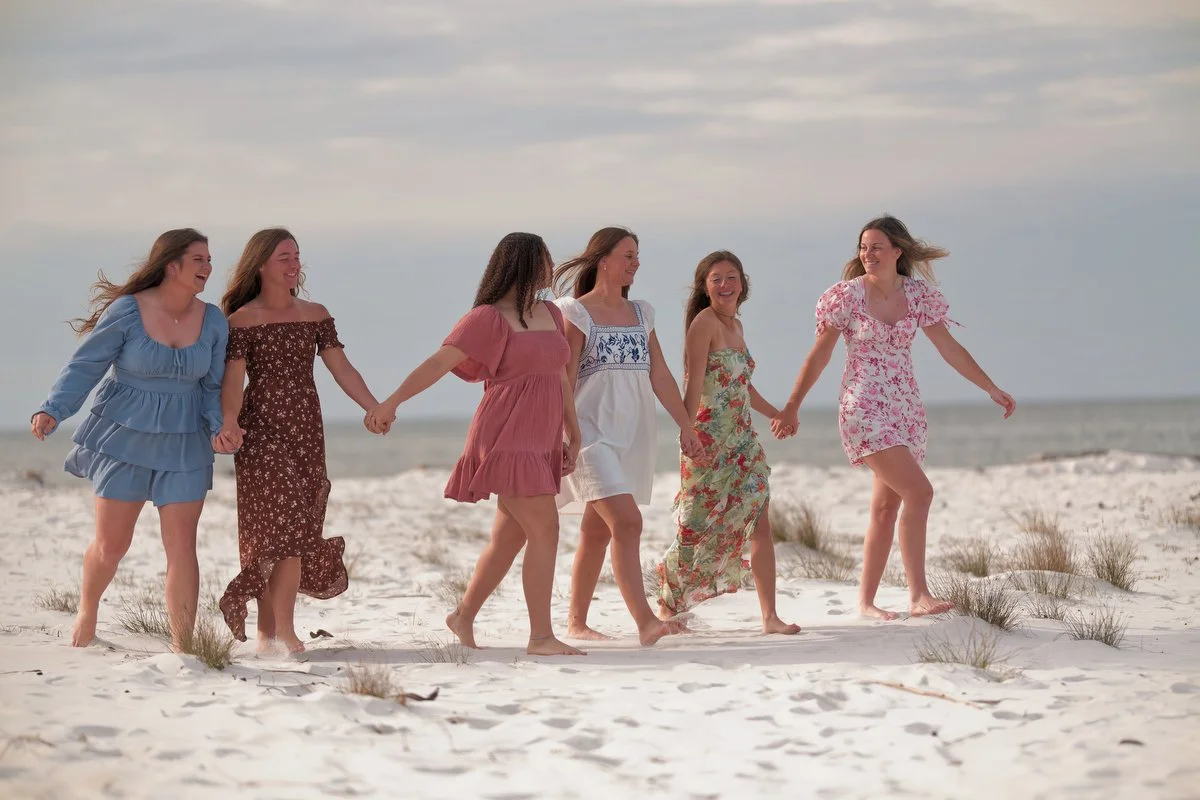 Six women holding hands, walking on a sandy beach with grass patches, smiling and enjoying a sunny day near the ocean.