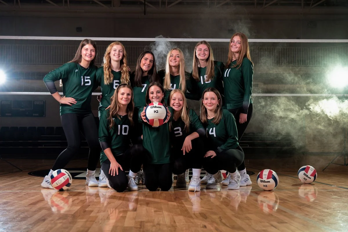 A girls' volleyball team poses in a gymnasium, wearing green uniforms. They are holding volleyballs and smiling for the camera, with a smoky ambiance and gym lights in the background.