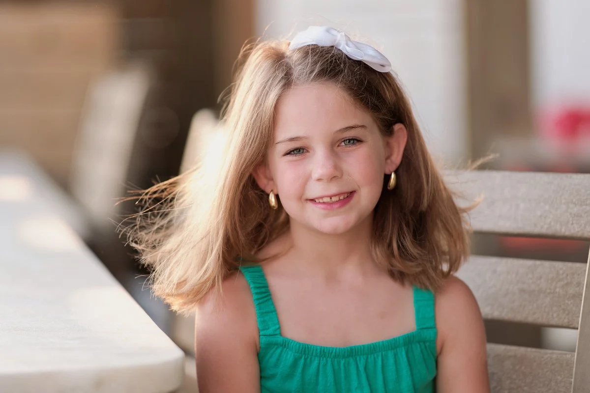 A young girl with light brown hair, wearing a white hair bow, gold hoop earrings, and a green dress, smiling outdoors.