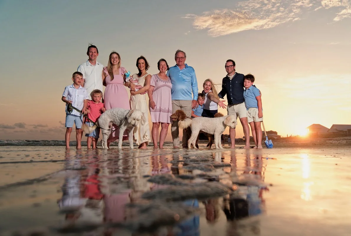Family group photo on the beach at sunset with dogs, including several children and adults, some holding dogs, with the reflective wet sand and ocean in the background in Fort Morgan, Alabama.