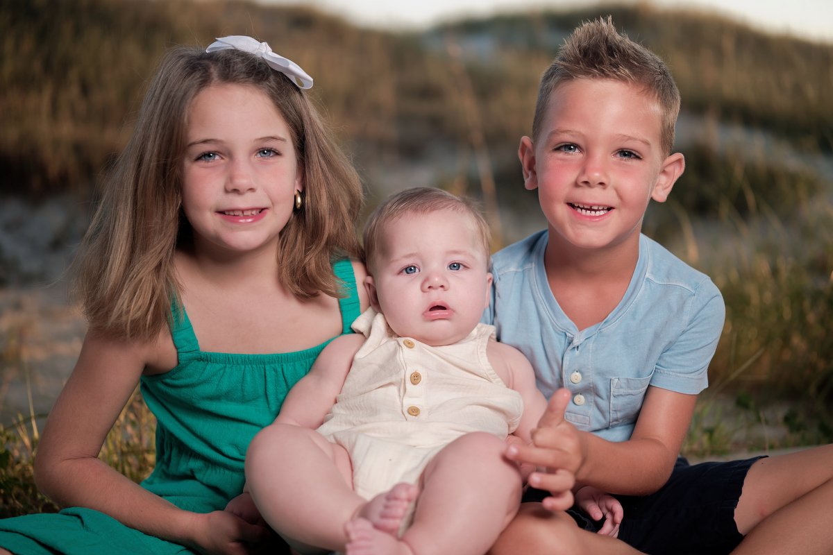Three young children, a girl and two boys, sitting outdoors on the grass near a body of water, smiling at the camera.