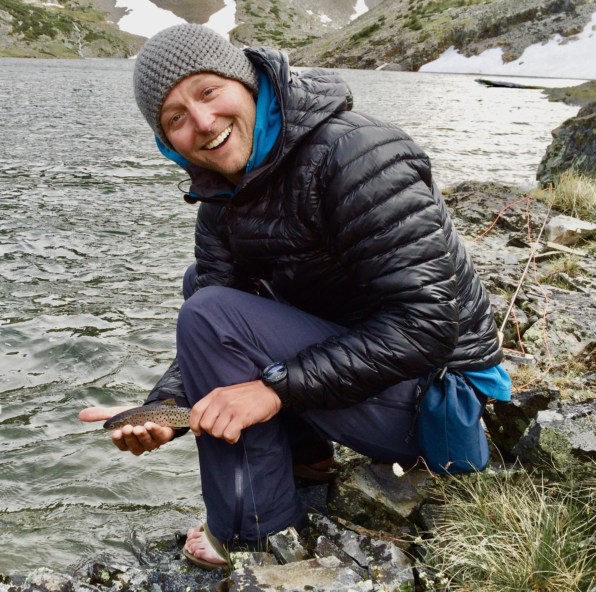 Man holds very small fish next to mountain lake