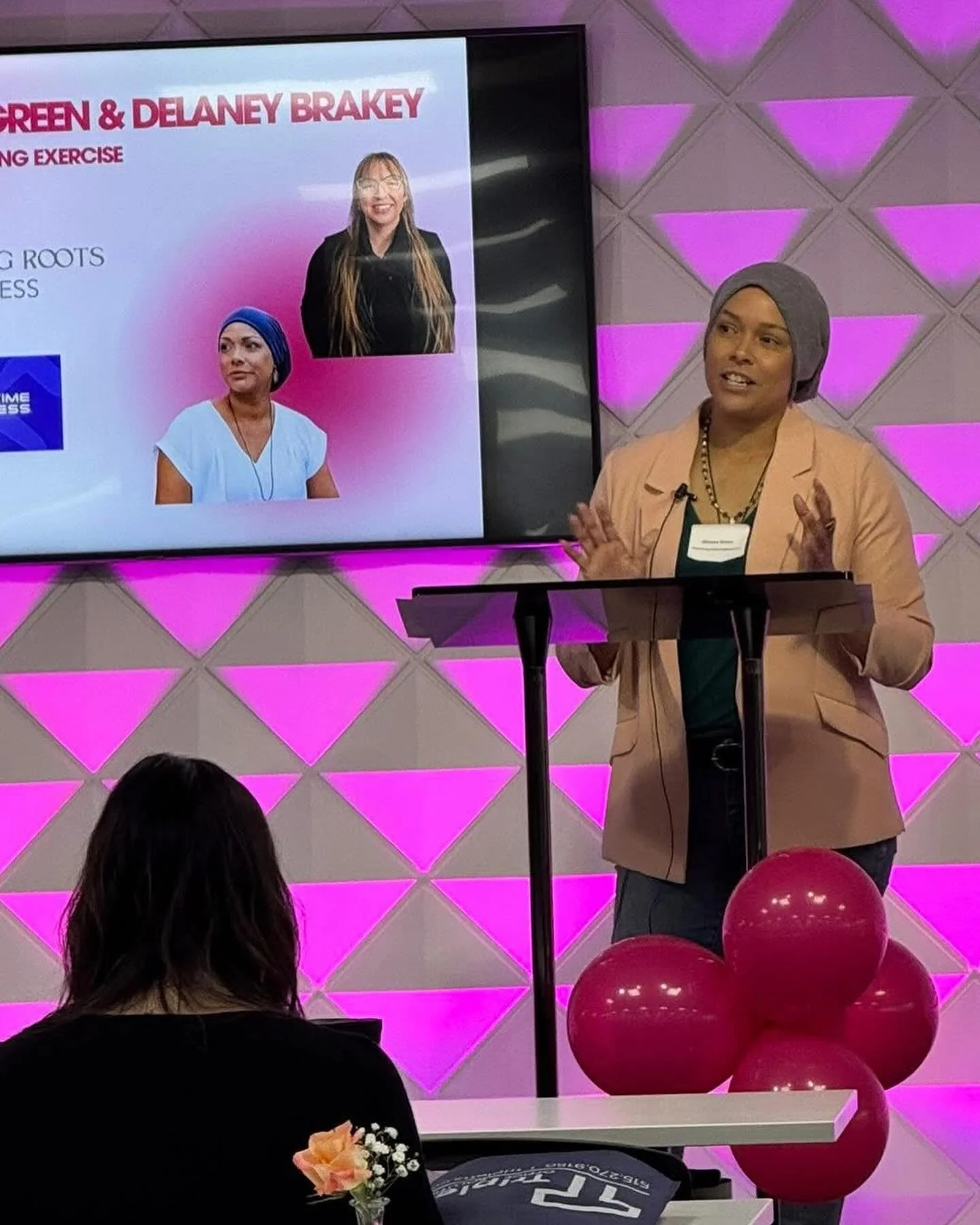 A woman is giving a presentation at a podium with a screen behind her displaying images of two women, one wearing a turban and the other with long straight hair. The room has a decorative wall with purple lighting and pink balloons on a table in the foreground.