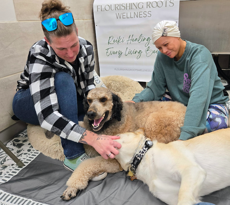 Two women and two dogs relaxing on a mat in front of a wellness sign, with one woman petting one of the dogs.