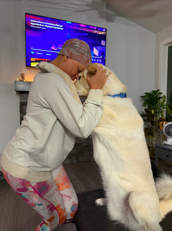 A person in a gray hoodie and colorful patterned leggings hugs a large white dog with a blue collar, both touching noses, in a cozy living room with a television and plants in the background.