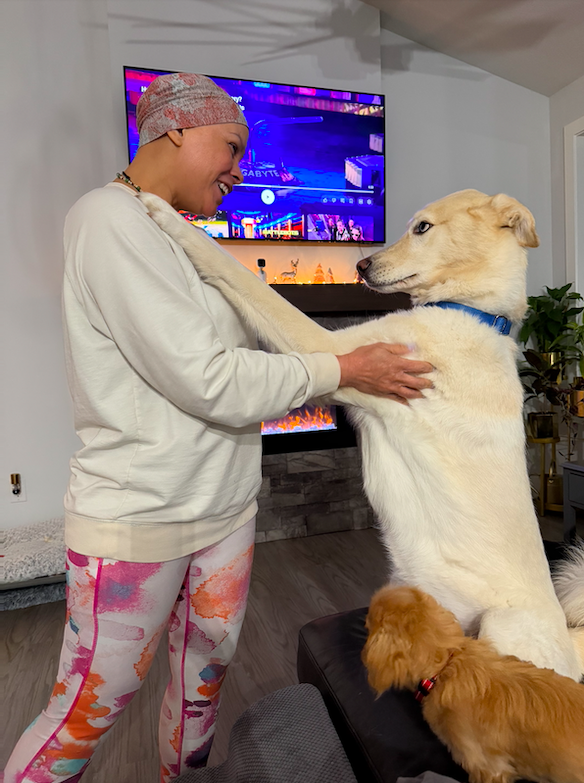 A woman in workout clothes and a headscarf smiling while holding a big white dog by the shoulders. Another smaller brown dog rests on a black couch nearby. The background includes a TV displaying a colorful scene and a cozy living room setting.