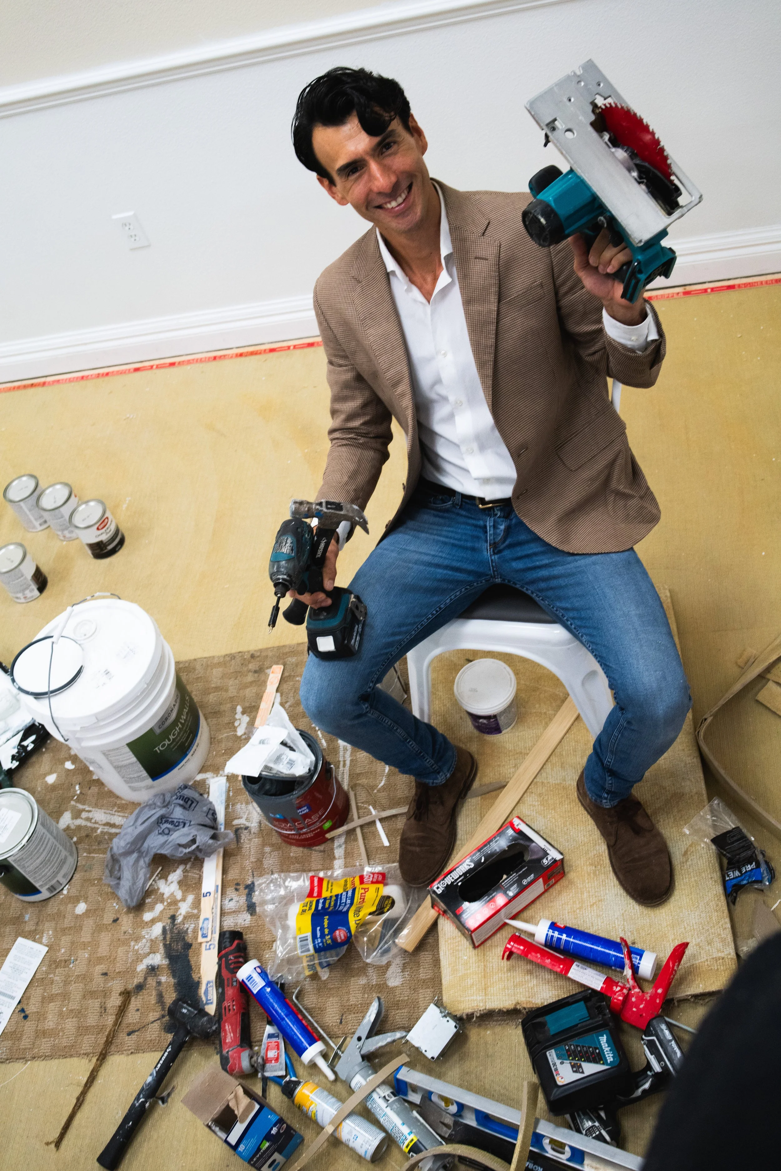 A man smiling and sitting on a white stool in a room with construction tools and supplies scattered around him, holding a power saw and drill.