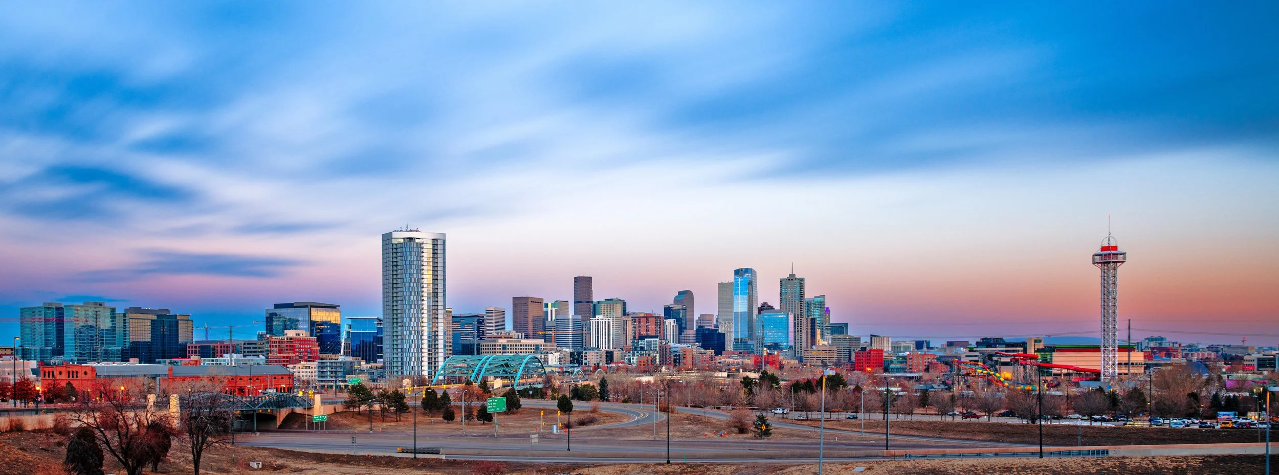 Skyline view of a city with tall buildings, a tower with a red top, and a colorful sunset sky.