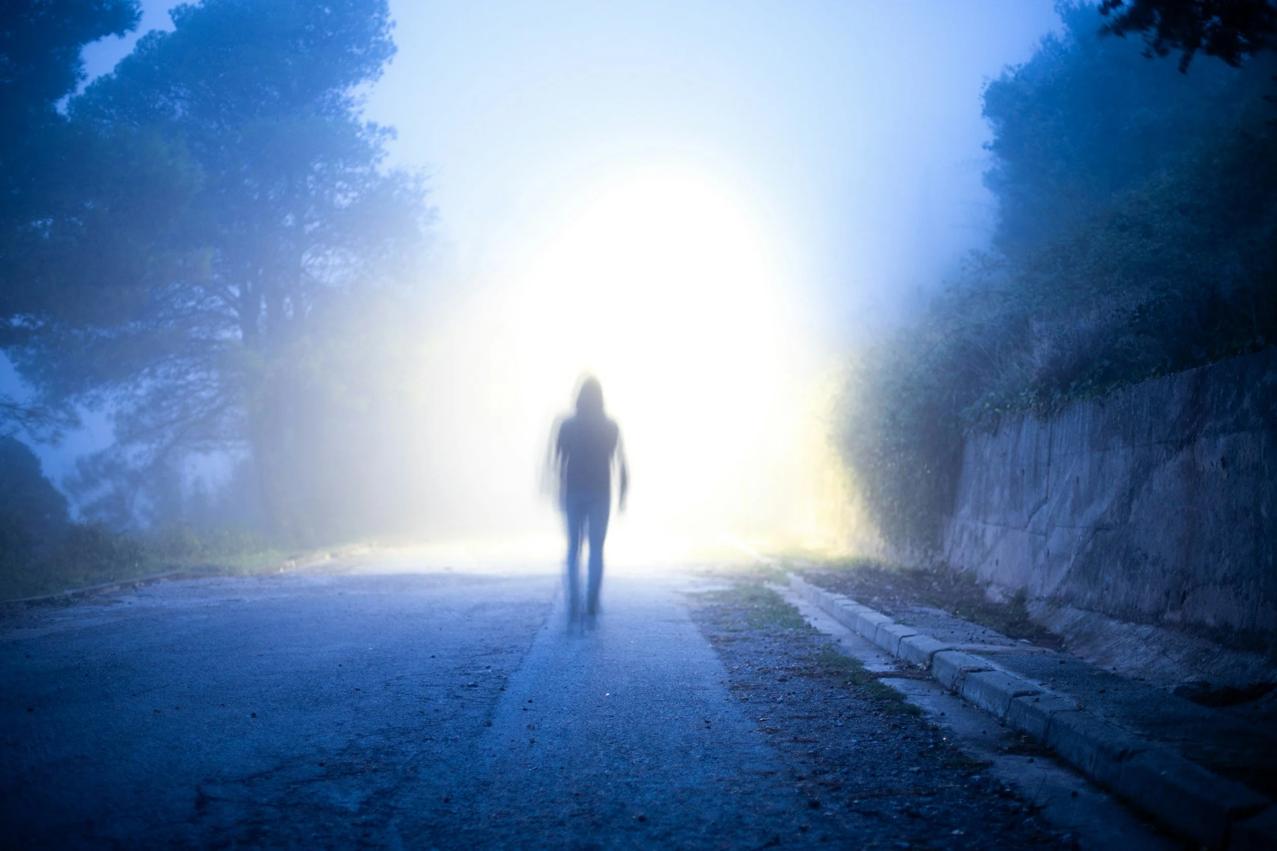 Silhouette floue d’une personne marchant sur une route brumeuse bordée d’arbres, baignée de lumière bleutée.