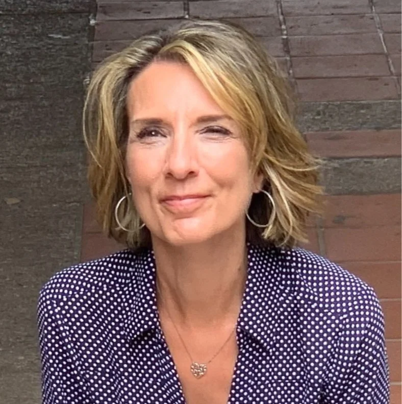 Smiling woman with short blonde hair wearing a polka dot blouse and hoop earrings.
