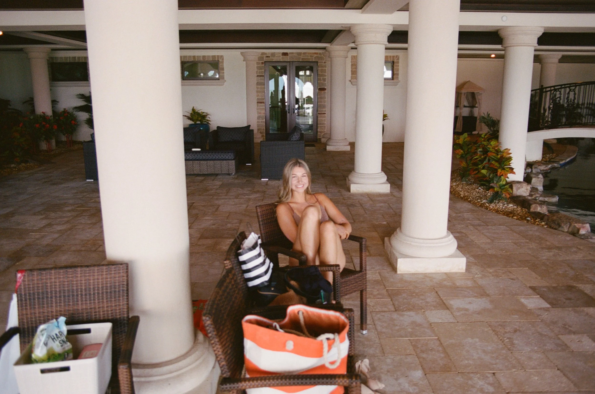 A woman sitting on a wicker chair on a covered outdoor patio area near a pond, surrounded by white columns, potted plants, and outdoor furniture.