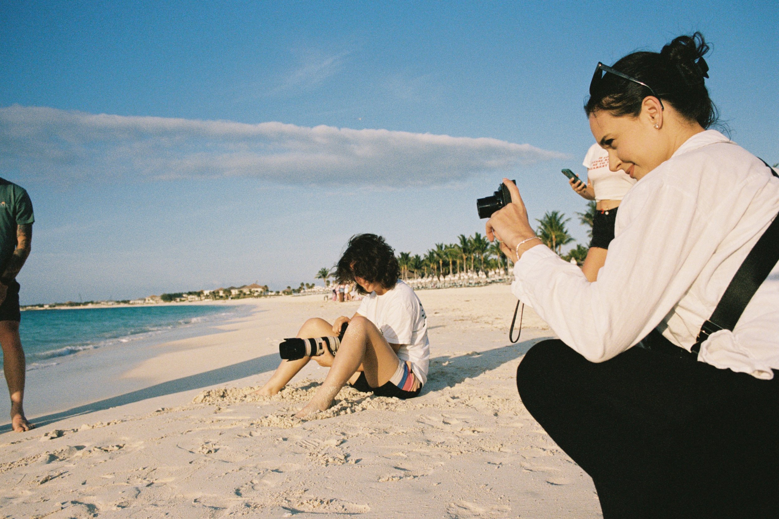 People taking photos on a sunny beach with sand, ocean waves, and palm trees in the background.