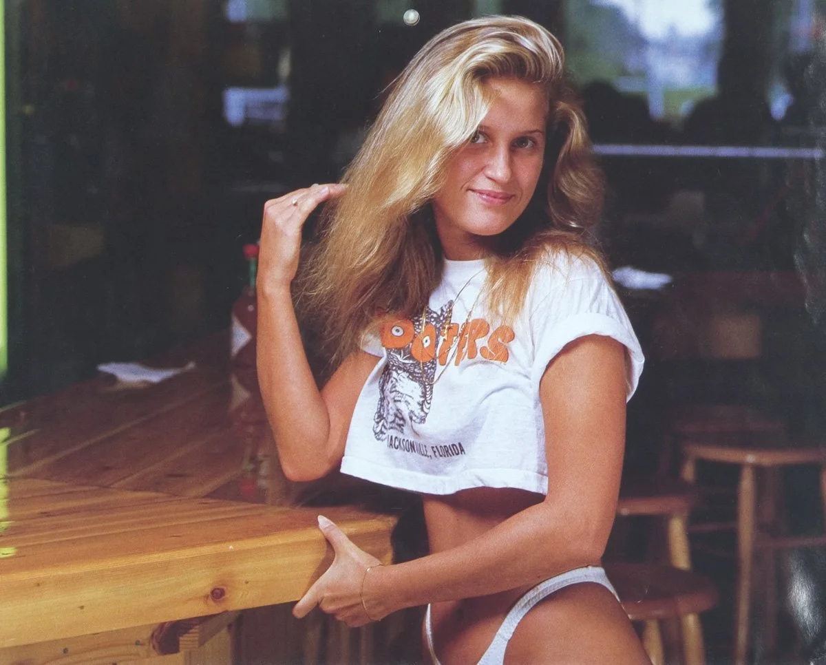 A Hooters Girl with long blonde hair sitting at a wooden table inside a restaurant or cafe, wearing a white crop top with orange text and a graphic, and holding her hair with one hand while smiling at the camera.