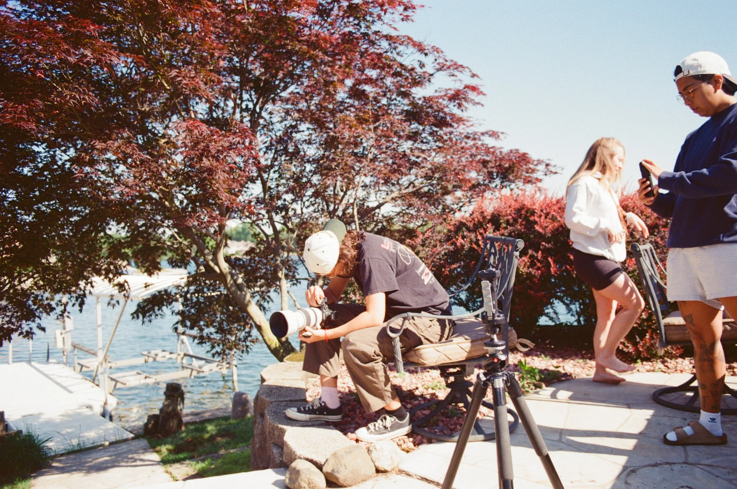 Three people near a body of water with trees and a dock, one person is sitting with a camera, and two others are standing and using phones.