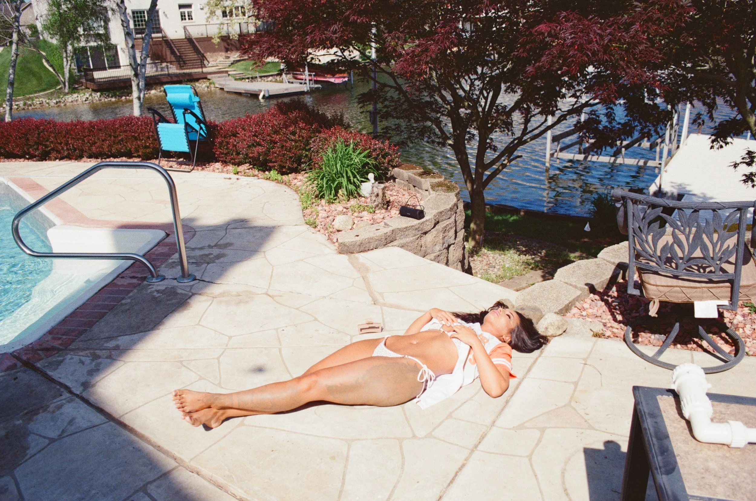A woman lying on a patio by a pool, sunbathing with a river and boats in the background, surrounded by trees and outdoor furniture.