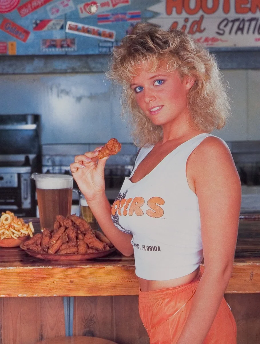 A Hooters Calendar Girl with curly blonde hair and blue eyes standing at a bar counter, holding a chicken wing. She is wearing a white sleeveless crop top with an orange logo and orange shorts.