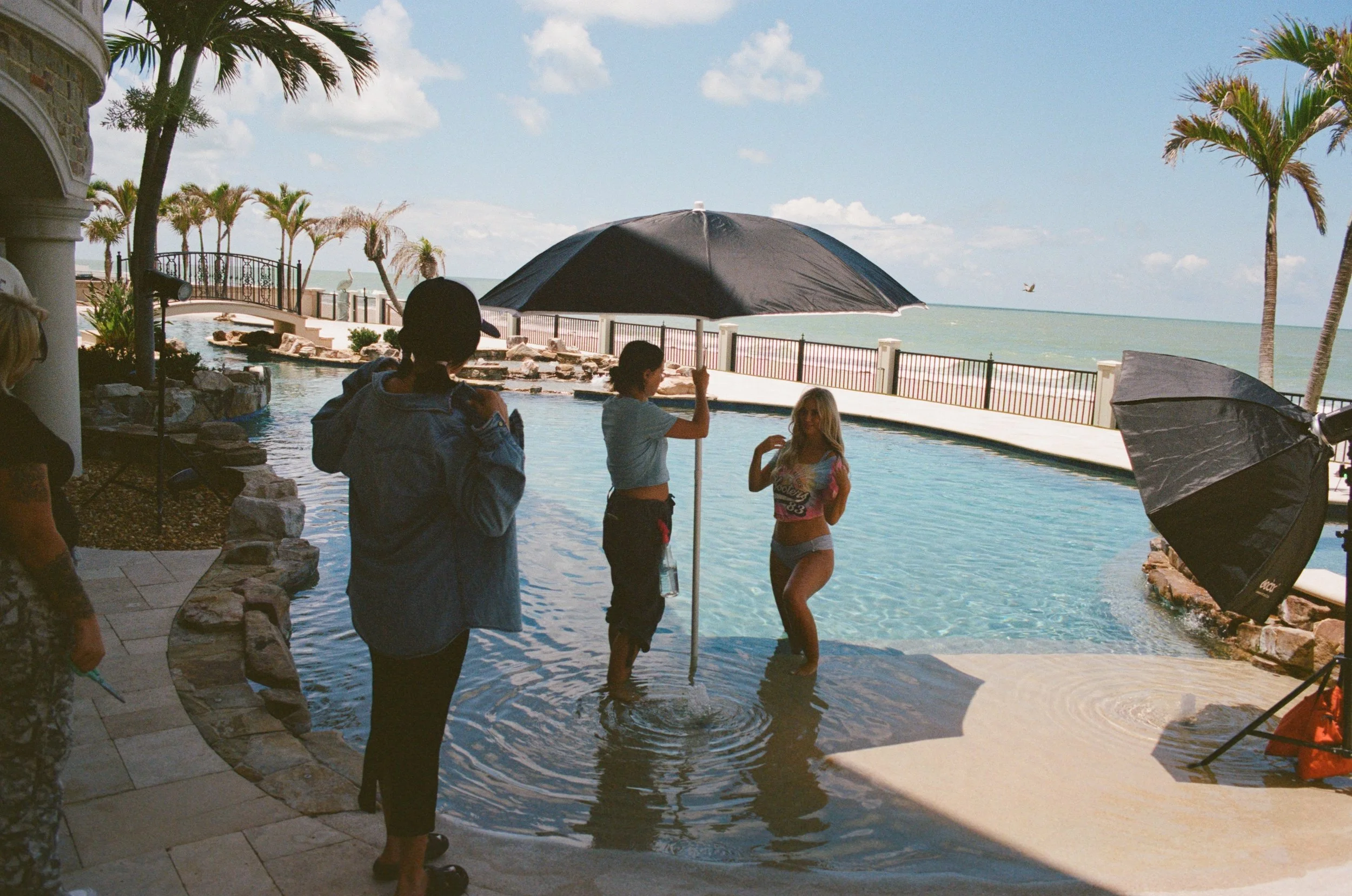 Beach photo shoot with a woman in swimwear, being photographed by a professional photographer near the ocean, with palm trees and umbrellas in the background.