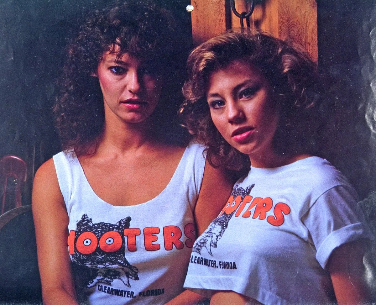 Two women with curly hair posing indoors against a wooden background, wearing matching T-shirts with the "Hooters" logo.