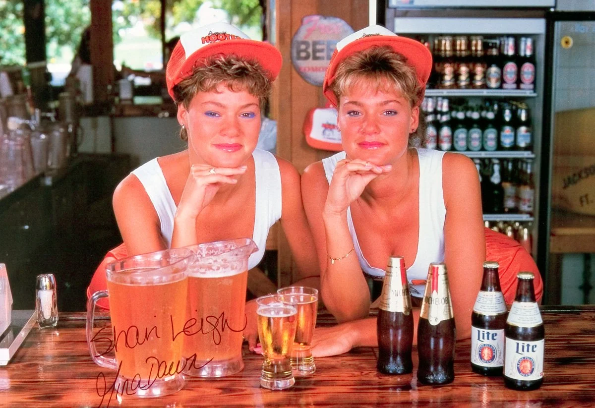 Two Hooters Calendar Girls in tank tops and hats sitting at a bar with beer bottles and glasses in front of them, inside a Hooters with beer bottles in the background.