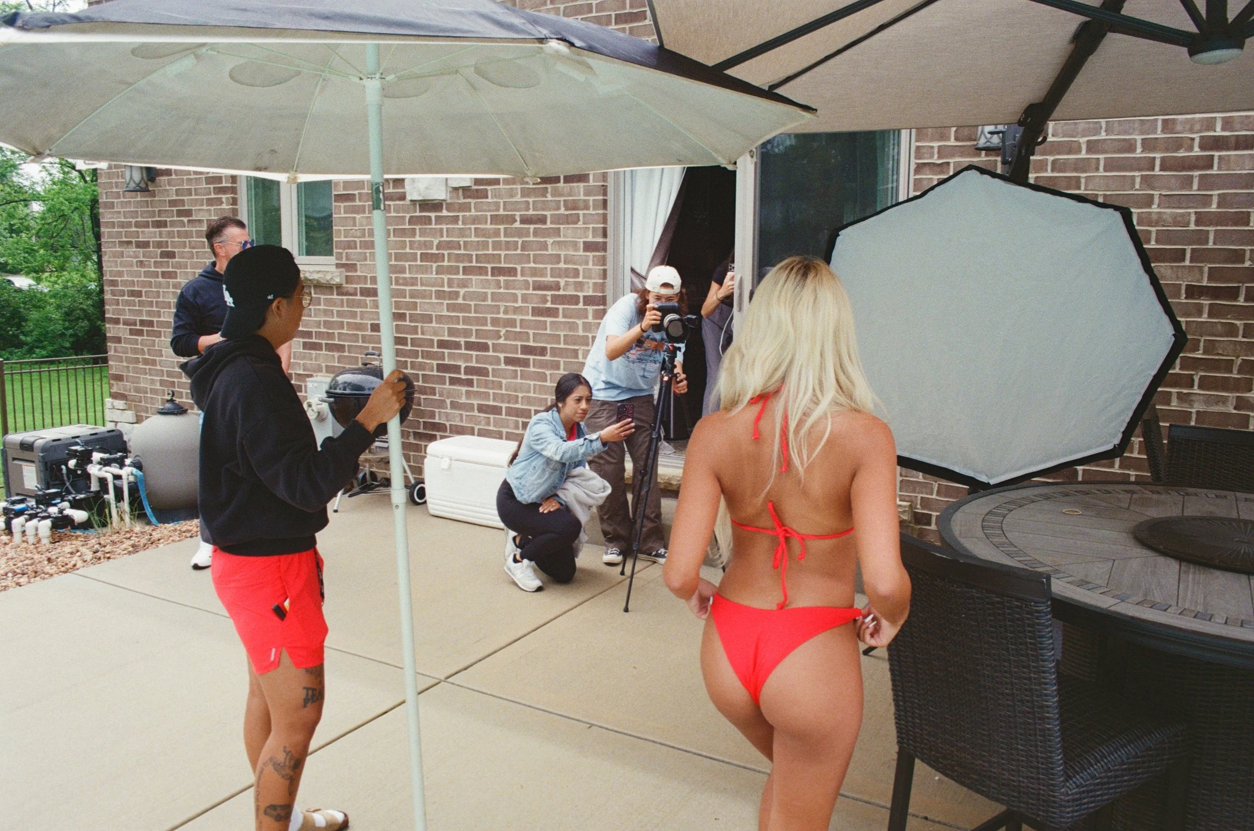 A woman in a red bikini is posing for a photo in an outdoor setting with multiple photographers and camera operators capturing her image. The scene is set on a patio with umbrellas and brick walls in the background.
